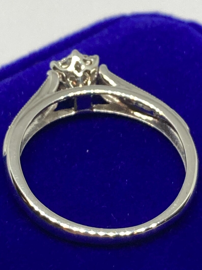 May include: A silver ring with a floral-inspired design at the center. The ring features a delicate band and a raised setting for the central stone. The ring is set against a blue background.