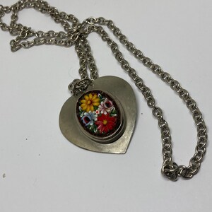 May include: Silver-toned necklace featuring a heart-shaped pendant. The pendant has a smaller oval inset with a floral mosaic design in red, yellow, blue, and white. The chain is a linked design.