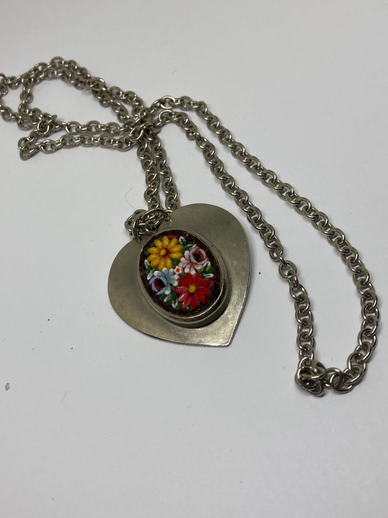 May include: Silver-toned necklace featuring a heart-shaped pendant. The pendant has a smaller oval inset with a floral mosaic design in red, yellow, blue, and white. The chain is a linked design.