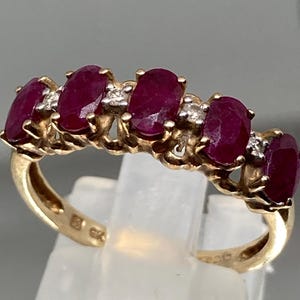 Peut inclure: Une bague en or ornée de cinq rubis rouges vifs de forme ovale. De petits diamants sont sertis entre les rubis. La bague est présentée sur un support transparent, mettant en valeur son design et l'agencement des pierres précieuses.