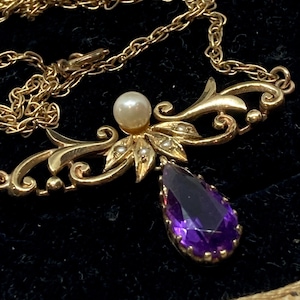 May include: A gold-toned necklace with a pendant featuring a pear-shaped, deep purple gemstone. The necklace has a delicate chain and is adorned with a pearl and small, seed-like pearls. The design includes ornate, swirling details.