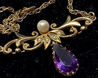 May include: A gold-toned necklace with a pendant featuring a pear-shaped, deep purple gemstone. The necklace has a delicate chain and is adorned with a pearl and small, seed-like pearls. The design includes ornate, swirling details.