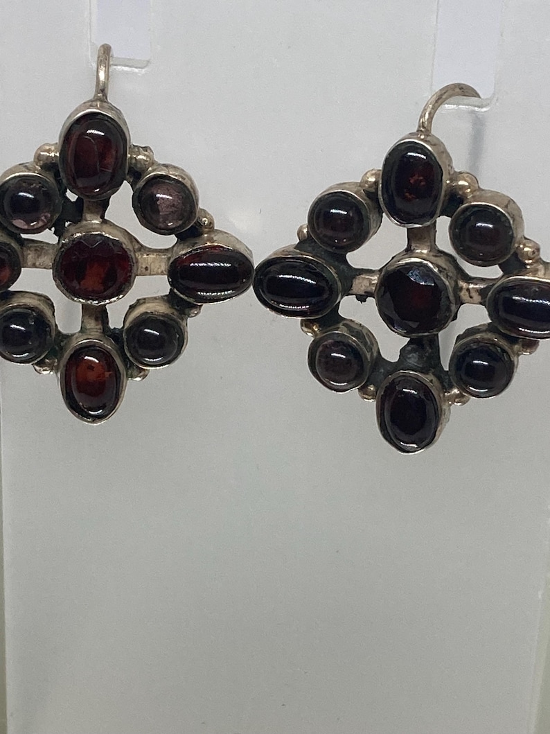 May include: A pair of silver earrings with a geometric design. Each earring features a central, round, dark red gemstone surrounded by smaller, oval-shaped gemstones of the same color. The earrings have a hook closure.