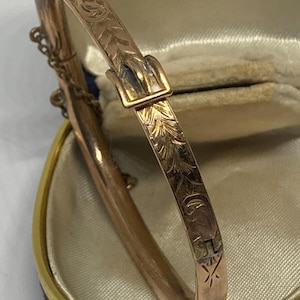 May include: A gold-toned metal bangle bracelet featuring an engraved leaf pattern and a buckle-style clasp. The bracelet is presented in a blue and cream-colored case with the text KOWLOON HONG KONG printed on the interior lining of the lid.