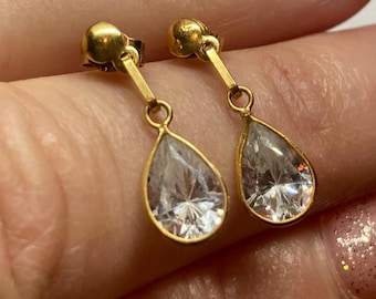 May include: A pair of gold-colored drop earrings. Each earring features a clear, teardrop-shaped stone set in a gold frame. The earrings are designed with a post and a small, round stud.
