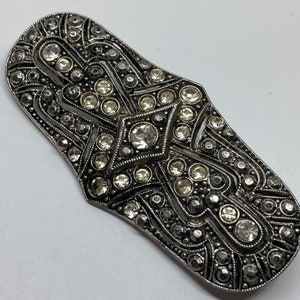 Puede incluir: Broche de estilo Art Déco con base de metal plateado. La broche rectangular con extremos redondeados está profusamente adornada con brillantes redondos y transparentes de varios tamaños. El diseño presenta patrones geométricos.