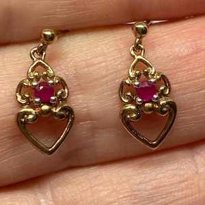 Puede incluir: Un par de pendientes dorados con un diseño en forma de corazón. Cada pendiente presenta una pequeña gema roja y redonda en el centro, rodeada por un patrón de filigrana decorativo. Los pendientes son de estilo colgante, con un poste para orejas perforadas.