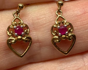 May include: A pair of gold-toned earrings with a heart-shaped design. Each earring features a small, round, red gemstone at the center, surrounded by a decorative filigree pattern. The earrings are dangling style, with a post for pierced ears.
