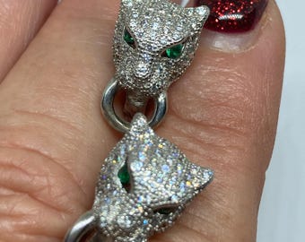 May include: A pair of silver earrings with a panther head design. The panther heads are encrusted with clear rhinestones and have green emerald eyes.