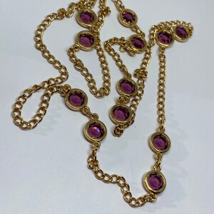 May include: A gold-toned necklace featuring a chain design with evenly spaced, round, purple gemstones. The gemstones are set in gold bezels, creating a contrast against the chain. The necklace is displayed on a white surface.