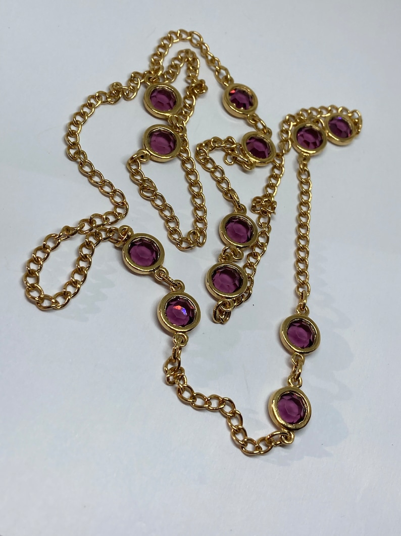 May include: A gold-toned necklace featuring a chain design with evenly spaced, round, purple gemstones. The gemstones are set in gold bezels, creating a contrast against the chain. The necklace is displayed on a white surface.