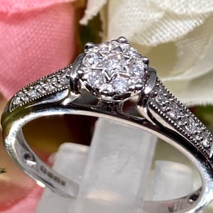 May include: A silver ring with a cluster of small, clear gemstones at the center, surrounded by smaller stones along the band. The ring is displayed on a clear stand, with a blurred floral background.