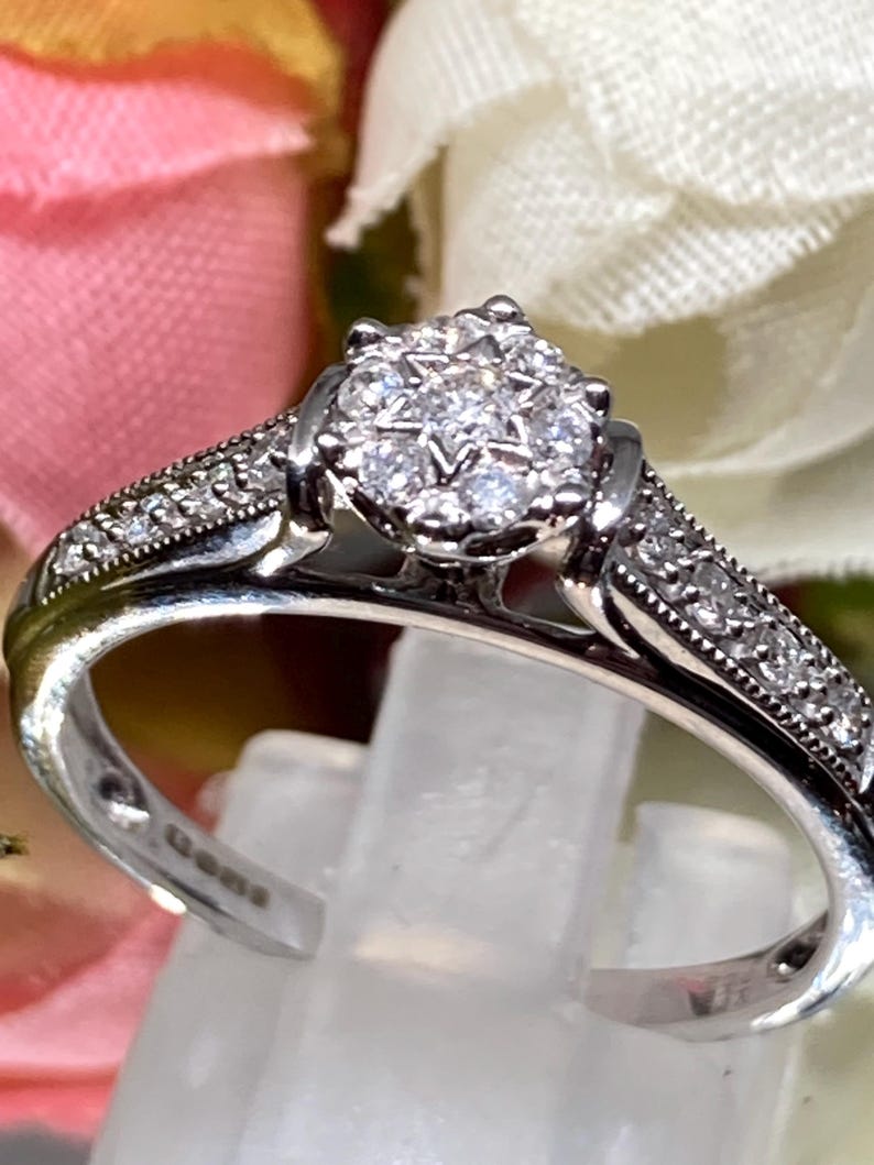 May include: A silver ring with a cluster of small, clear gemstones at the center, surrounded by smaller stones along the band. The ring is displayed on a clear stand, with a blurred floral background.