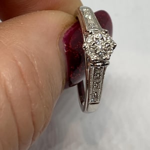 May include: A silver ring with a cluster of small, round, clear stones at the center. The band is also adorned with smaller stones. The ring is held up against a blurred background.
