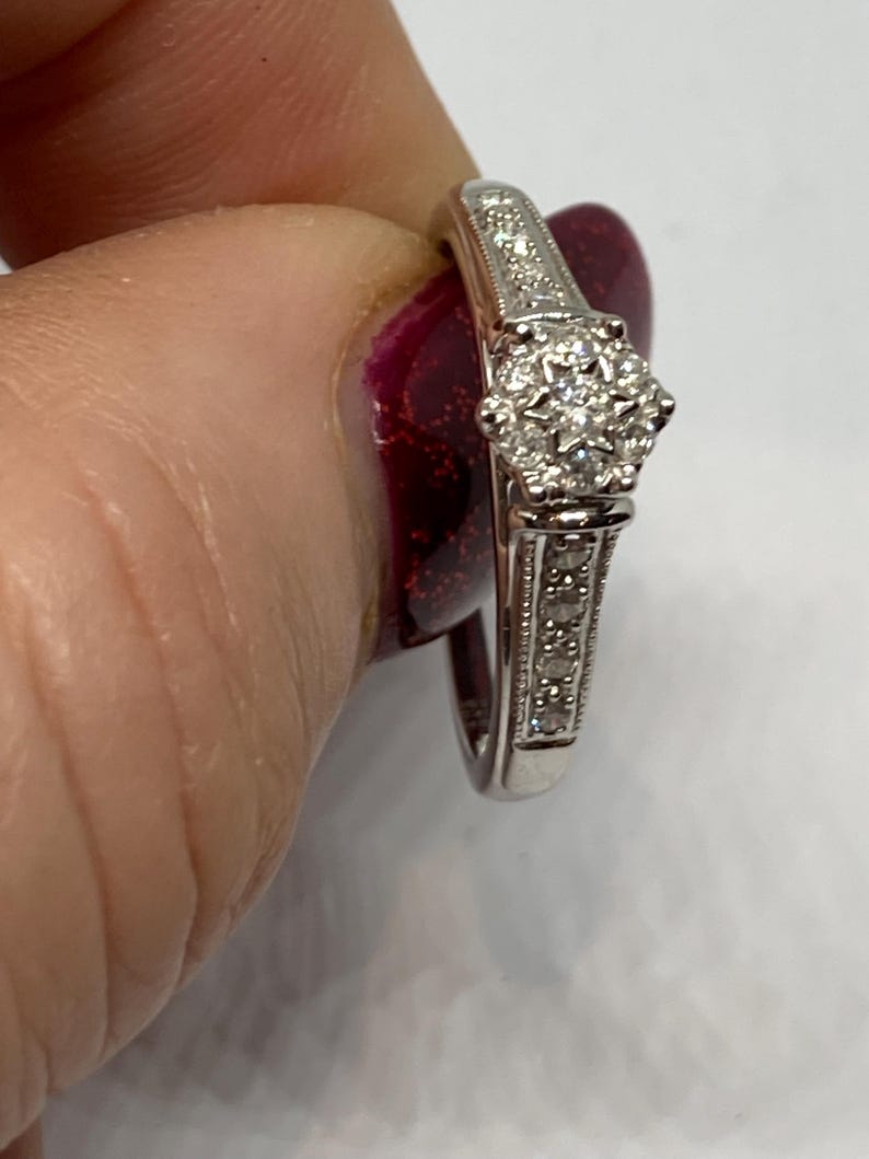 May include: A silver ring with a cluster of small, round, clear stones at the center. The band is also adorned with smaller stones. The ring is held up against a blurred background.
