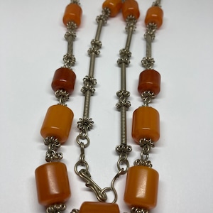 May include: A vintage necklace featuring amber-colored barrel beads strung on silver-toned metal wire. The beads vary in shade from light orange to deep amber. The necklace has a clasp closure and decorative metal accents.