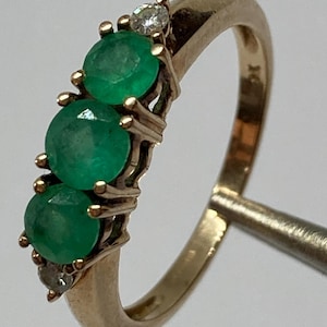 Puede incluir: Un anillo de oro con tres esmeraldas verdes redondas y dos pequeños diamantes. Las piedras preciosas están engastadas en un diseño clásico y elegante, que resalta el atractivo atemporal del anillo. Una pieza de joyería fina.