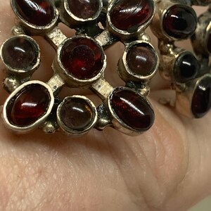 May include: Close-up of vintage silver-toned earrings featuring oval-shaped, deep red gemstones. The gemstones are set in a grid-like pattern, creating a striking contrast against the metal. The earrings are likely antique or vintage.