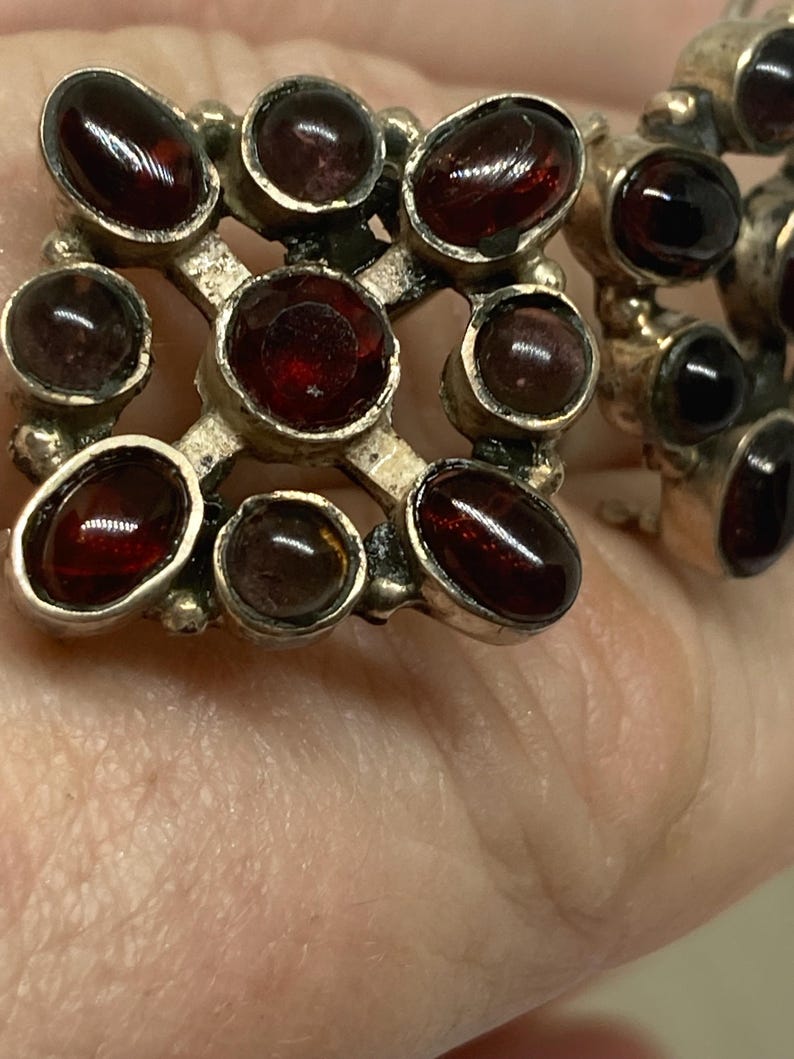 May include: Close-up of vintage silver-toned earrings featuring oval-shaped, deep red gemstones. The gemstones are set in a grid-like pattern, creating a striking contrast against the metal. The earrings are likely antique or vintage.