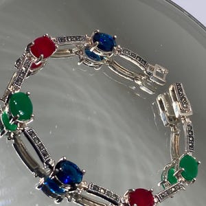 May include: Silver bracelet featuring oval-cut gemstones in red, blue, and green. The bracelet has a link design with marcasite accents. The clasp is visible, and the bracelet is displayed on a reflective surface.