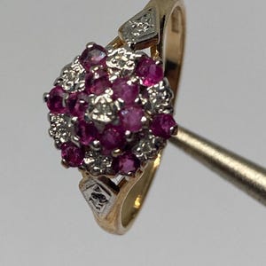 May include: A gold ring featuring a cluster head set with multiple round-cut pinkish-red rubies and small clear accent stones. The band has a polished gold finish with decorative silver-toned metal accents on the shoulders near the central setting.