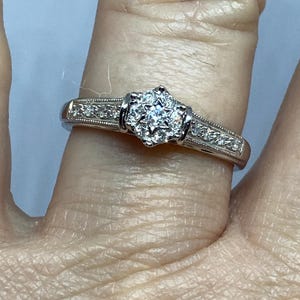 May include: A silver ring with a cluster of small, sparkling stones at the center. The band features smaller stones and delicate detailing. The ring is set on a finger, showcasing its design and craftsmanship.