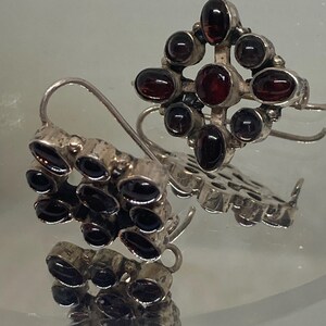 May include: A pair of silver earrings with dark red gemstones. One earring features a cluster of stones, while the other has a cross-shaped design with a central stone and surrounding gems. The earrings have a hook closure.