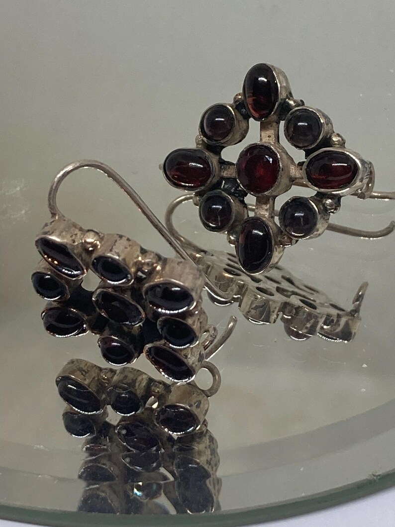 May include: A pair of silver earrings with dark red gemstones. One earring features a cluster of stones, while the other has a cross-shaped design with a central stone and surrounding gems. The earrings have a hook closure.