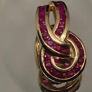 May include: A gold pendant featuring a swirling design, set with numerous small, round, deep red gemstones. The pendant's shape resembles an infinity symbol, with the gemstones closely set together.