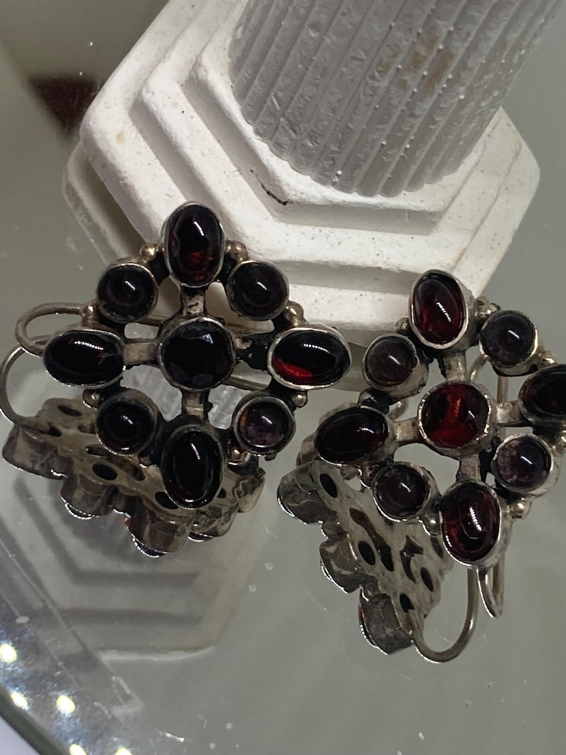 May include: A pair of silver-toned earrings featuring a cluster of dark red and black oval-shaped gemstones. The earrings have a floral design at the base and a hook closure. The gemstones are set in a silver metal frame.