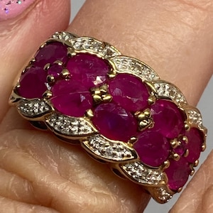 May include: A gold ring featuring a cluster of oval-cut deep pink rubies arranged in a multi-row pattern. The band is accented with small, clear pavé-set stones along the edges, creating a textured, layered appearance across the surface of the ring.