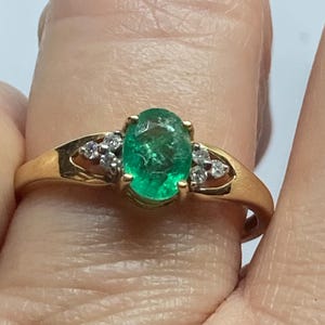 May include: A gold ring with an oval emerald gemstone at its centre. The ring is accented with small, round diamonds on either side of the emerald. The band has a simple, elegant design.