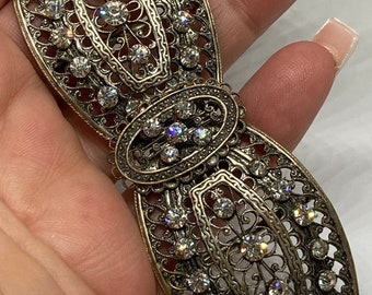 May include: An antique silver-toned filigree brooch with clear rhinestones. The brooch has an oval center and two elongated, openwork sections, all adorned with sparkling stones. The intricate design and vintage style make it a unique accessory.