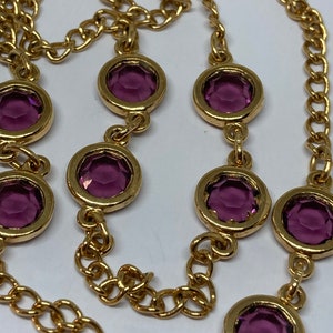 May include: A gold-toned necklace featuring a chain design with alternating round, bezel-set, purple gemstones. The gemstones are evenly spaced along the chain, creating a continuous pattern. The necklace is displayed against a white background.