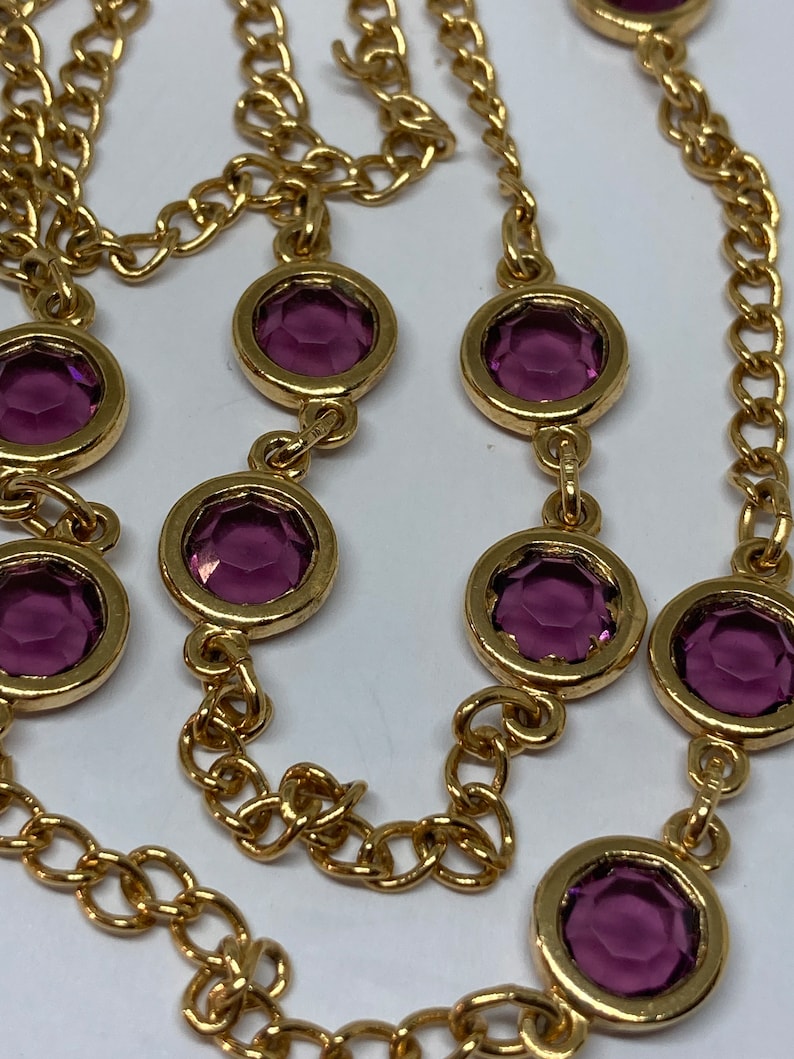 May include: A gold-toned necklace featuring a chain design with alternating round, bezel-set, purple gemstones. The gemstones are evenly spaced along the chain, creating a continuous pattern. The necklace is displayed against a white background.