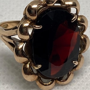 May include: A gold ring featuring a large, oval-cut, deep red gemstone set in the center. The stone is surrounded by a decorative, scalloped gold frame with rounded edges. The band of the ring is made of polished gold and has a split-shank design.