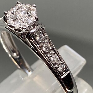 May include: A silver ring with a cluster of small diamonds at the center. The band features a row of smaller diamonds and detailed metalwork. The ring is set on a clear display stand, showcasing its design.