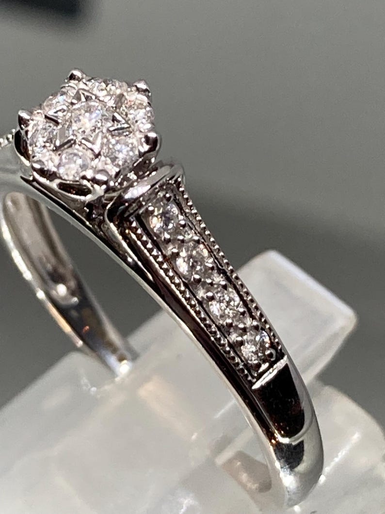 May include: A silver ring with a cluster of small diamonds at the center. The band features a row of smaller diamonds and detailed metalwork. The ring is set on a clear display stand, showcasing its design.