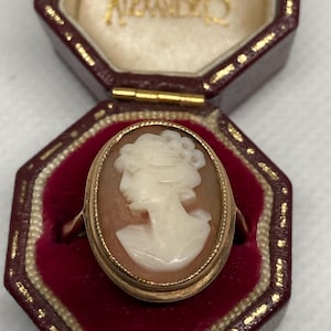 May include: An oval cameo ring featuring a white carved profile of a person against a light brown background, set in a gold-toned metal frame. The ring is displayed inside an octagonal, dark red velvet-lined jewelry box with gold lettering.