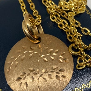 May include: A gold-colored necklace featuring a round pendant with a tree design. The pendant has a textured, matte finish. The chain is a classic link style, and the necklace is displayed on a dark blue surface.