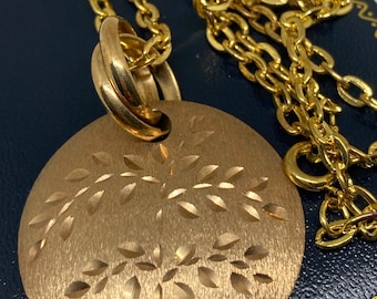 May include: A gold-colored necklace featuring a round pendant with a tree design. The pendant has a textured, matte finish. The chain is a classic link style, and the necklace is displayed on a dark blue surface.