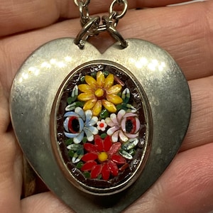 May include: Silver-toned heart pendant necklace with a mosaic floral design. The pendant features a heart-shaped base with an oval inset showcasing a colorful arrangement of flowers in red, yellow, blue, and pink. The necklace has a chain.