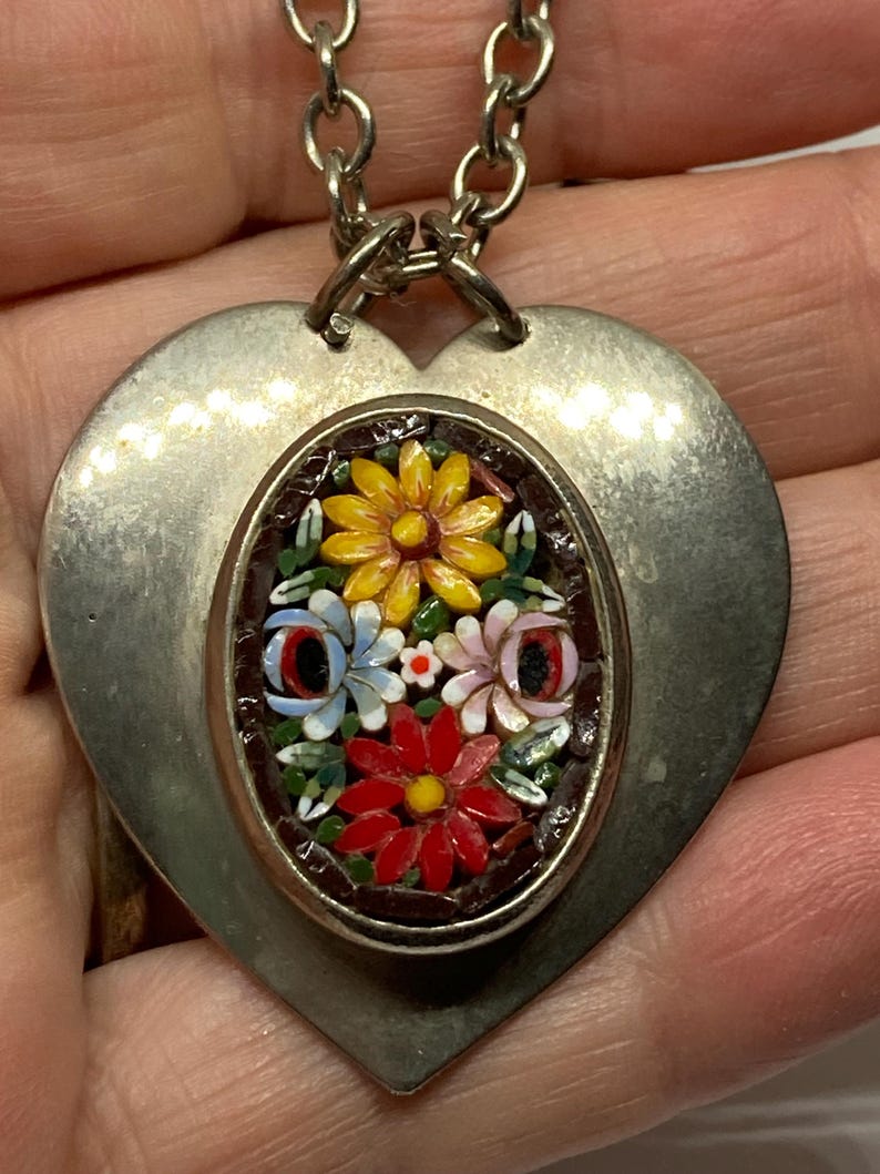May include: Silver-toned heart pendant necklace with a mosaic floral design. The pendant features a heart-shaped base with an oval inset showcasing a colorful arrangement of flowers in red, yellow, blue, and pink. The necklace has a chain.