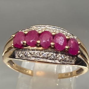 May include: A gold ring with five oval rubies set in a row, with a band of small diamonds above and below. The ring has a polished gold band and is displayed on a clear stand. The ring is likely made of 9ct gold.