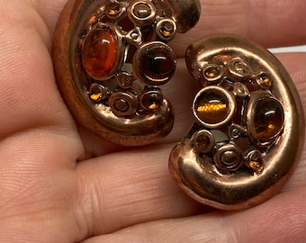 May include: A pair of copper-toned earrings with a crescent shape. Each earring features amber-colored, round, and oval-shaped stones and decorative swirls. The earrings are displayed on a light background.