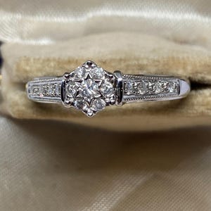 May include: A silver ring with a cluster of small diamonds in a starburst design at the center. The band is adorned with smaller diamonds and intricate detailing. The ring is set against a neutral background.