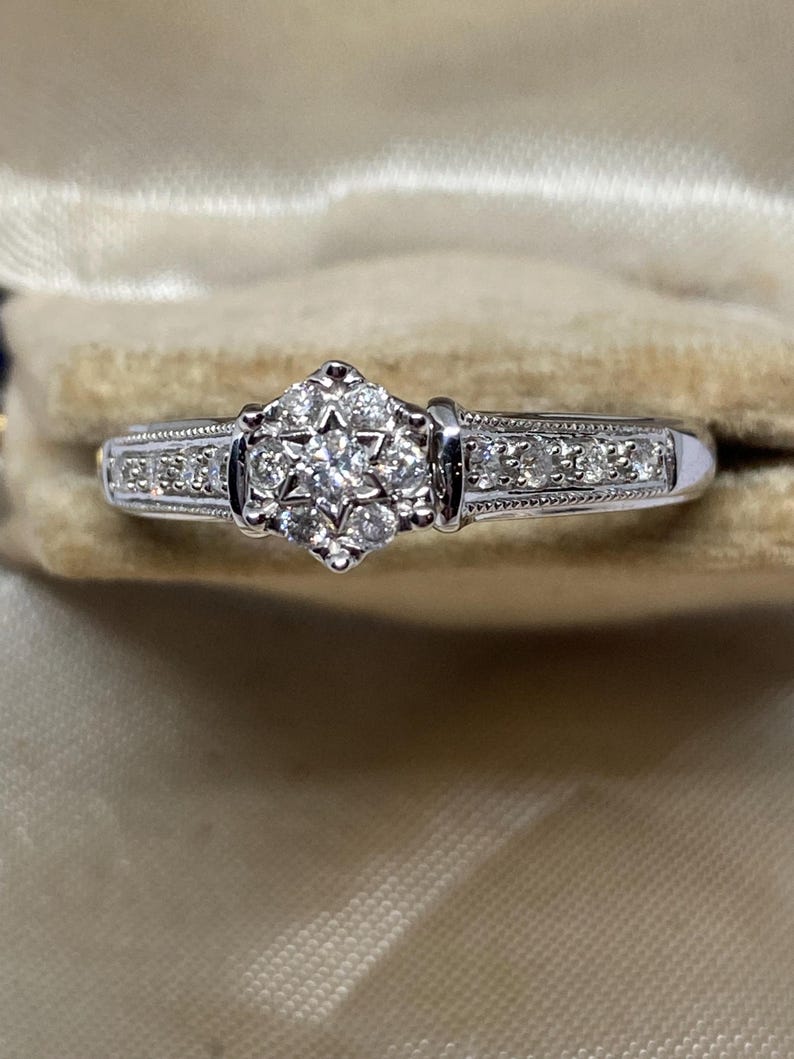 May include: A silver ring with a cluster of small diamonds in a starburst design at the center. The band is adorned with smaller diamonds and intricate detailing. The ring is set against a neutral background.
