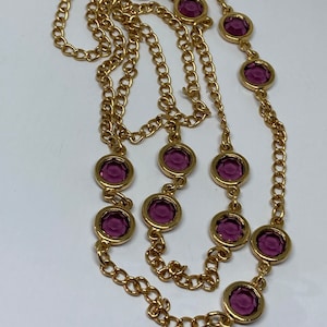 May include: A gold-toned necklace featuring a series of round, deep purple gemstones set in gold bezels. The chain is a classic link style, creating a layered effect. The necklace is displayed against a white background.