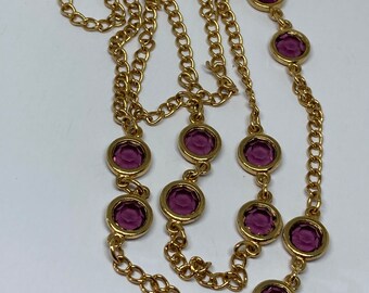 May include: A gold-toned necklace featuring a series of round, deep purple gemstones set in gold bezels. The chain is a classic link style, creating a layered effect. The necklace is displayed against a white background.