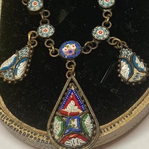 May include: Vintage mosaic necklace with a silver chain. The necklace features a central teardrop pendant and smaller floral and geometric mosaic designs in shades of blue, red, white, and green. The necklace is displayed on a black surface.